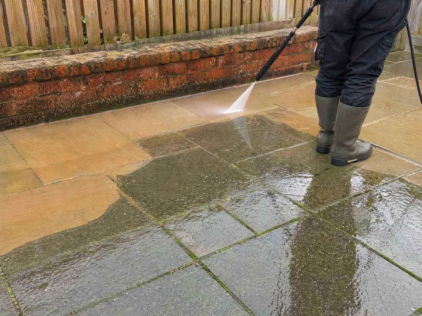 Pressure washing a paved patio beside a brick garden wall, the cleaned slabs next to ones still grey with moss